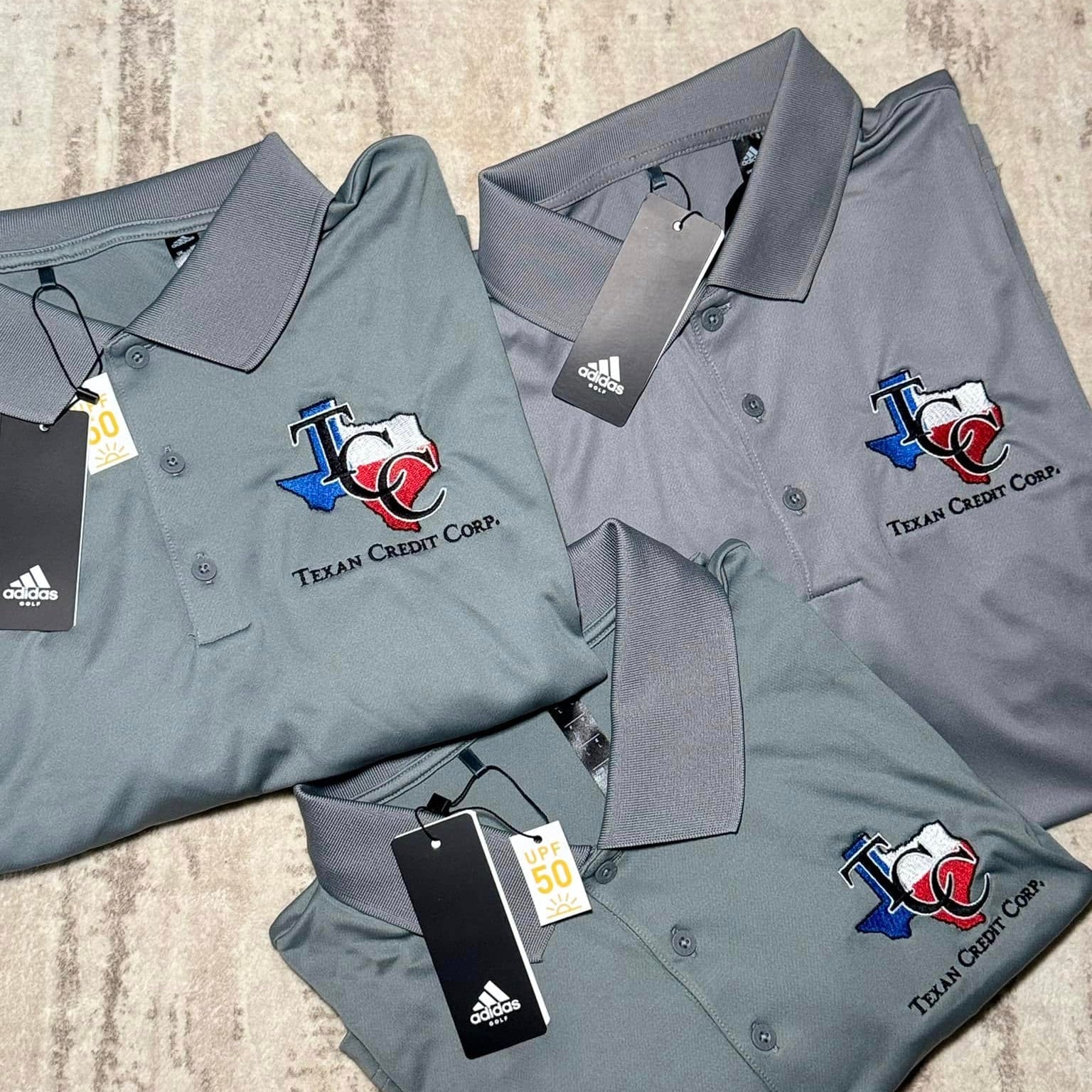 Three gray polo shirts with Texas Credit Corp logo and Adidas tags on a textured surface.