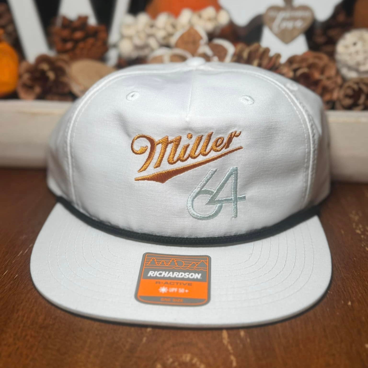 White cap with 'Miller 64' embroidered branding on a wooden surface with decorative items in the background