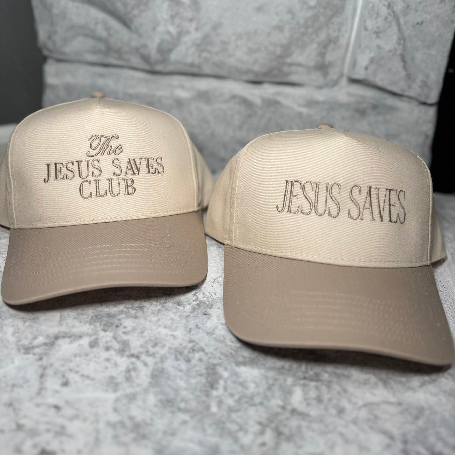 Two beige baseball caps with Jesus Saved Club on a stone surface